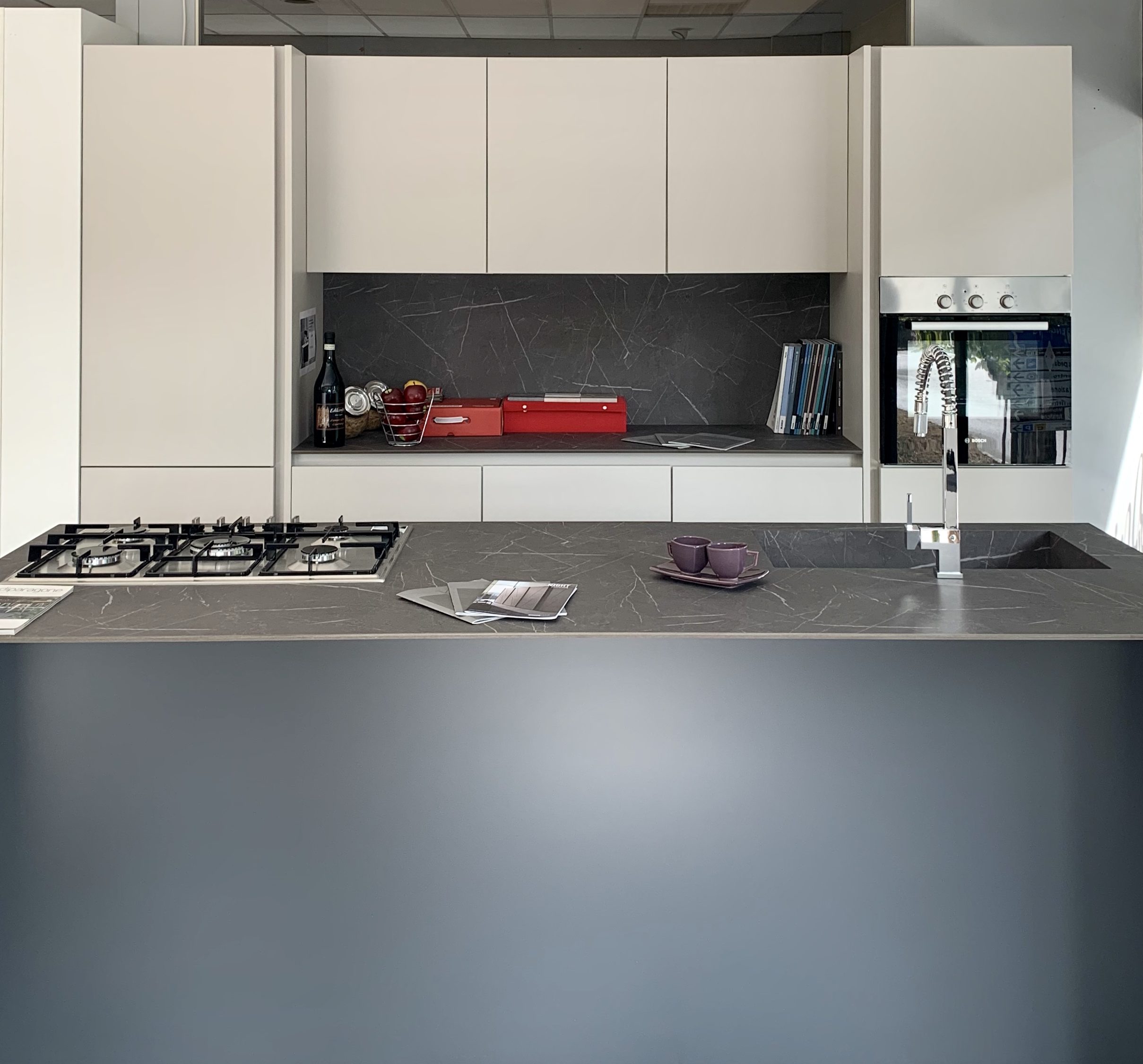 Immagine raffigurante una cucina spagnolcucine. Da destra a sinistra si visualizza un lavandino un piano cottura e delle tazzine. Sullo sfondo un mobile con un forno, libri, bottiglie e una fruttiera